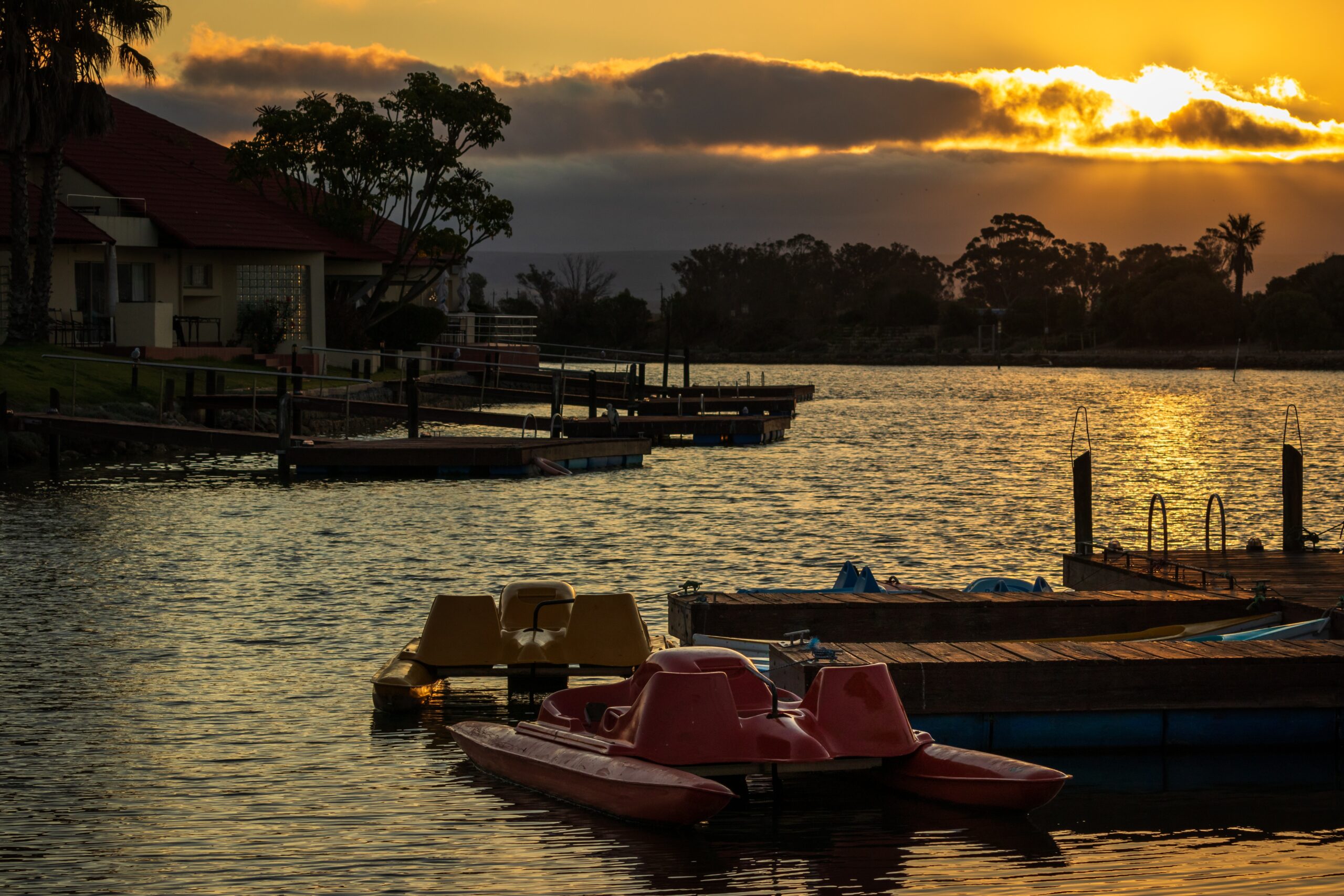 Sunset view of serene lake with pedal boats moored at wooden dock, silhouetted trees and houses in the background