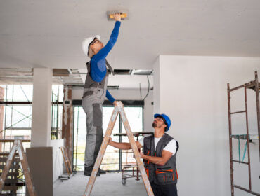 Two men installing fixtures on a ceiling in a room, using tools and ladders for their work